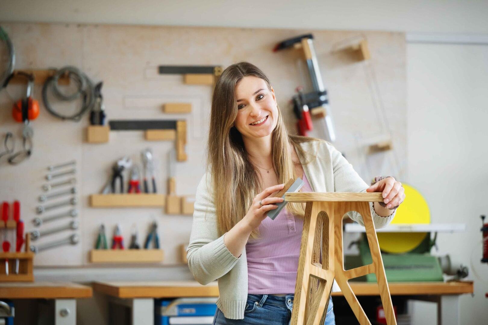 Eine Frau lächelt, während sie in einer Werkstatt einen Holzschemel abschleift. Werkzeuge und Geräte sind ordentlich an der Wand hinter ihr angeordnet.