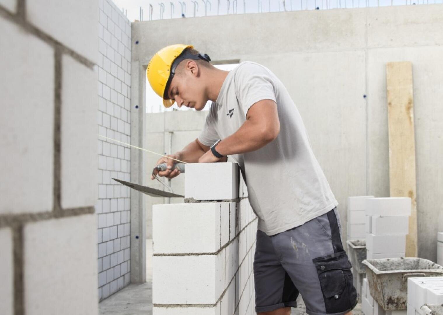 Ein Bauarbeiter, der einen gelben Schutzhelm und ein graues Hemd trägt, verlegt mit einer Kelle weiße Ziegelsteine an einer Wand auf einer Baustelle.