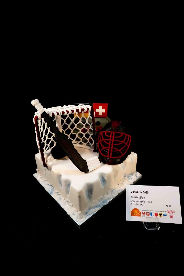 Ein Kuchen, verziert mit einem Eishockeytor, einem Schläger, einem Puck, einem Helm und einer Schweizer Flagge obenauf, platziert auf einem Sockel im Eis-Design, daneben eine Karte mit Angaben zum Hersteller.