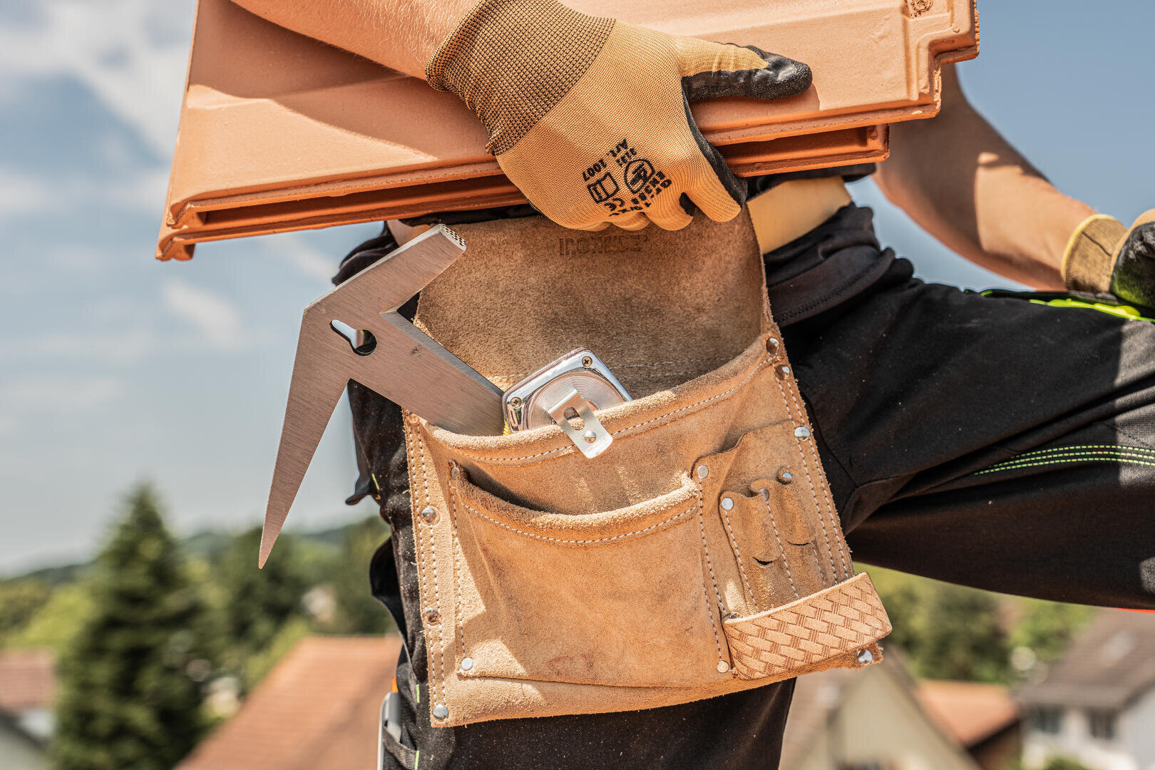 Nahaufnahme des Werkzeuggürtels eines Bauarbeiters mit einem Winkel, einem Maßband und Bleistiften. Der Arbeiter trägt Handschuhe und hält Dachziegel, während Häuser und Bäume im Hintergrund verschwimmen.