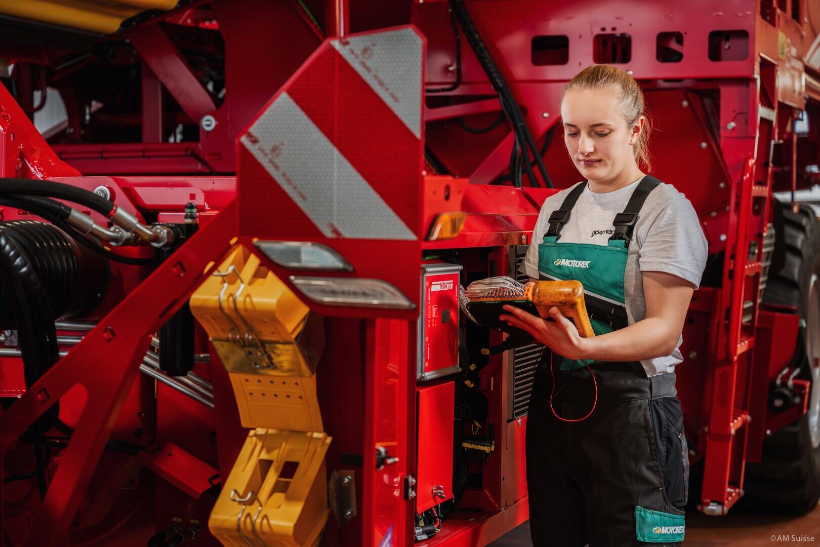 Eine Frau in einem Arbeitsoverall steht neben einer großen roten Maschine, prüft Kabel und macht sich Notizen auf einem Klemmbrett oder einem Tablet in einer industriellen oder landwirtschaftlichen Umgebung.