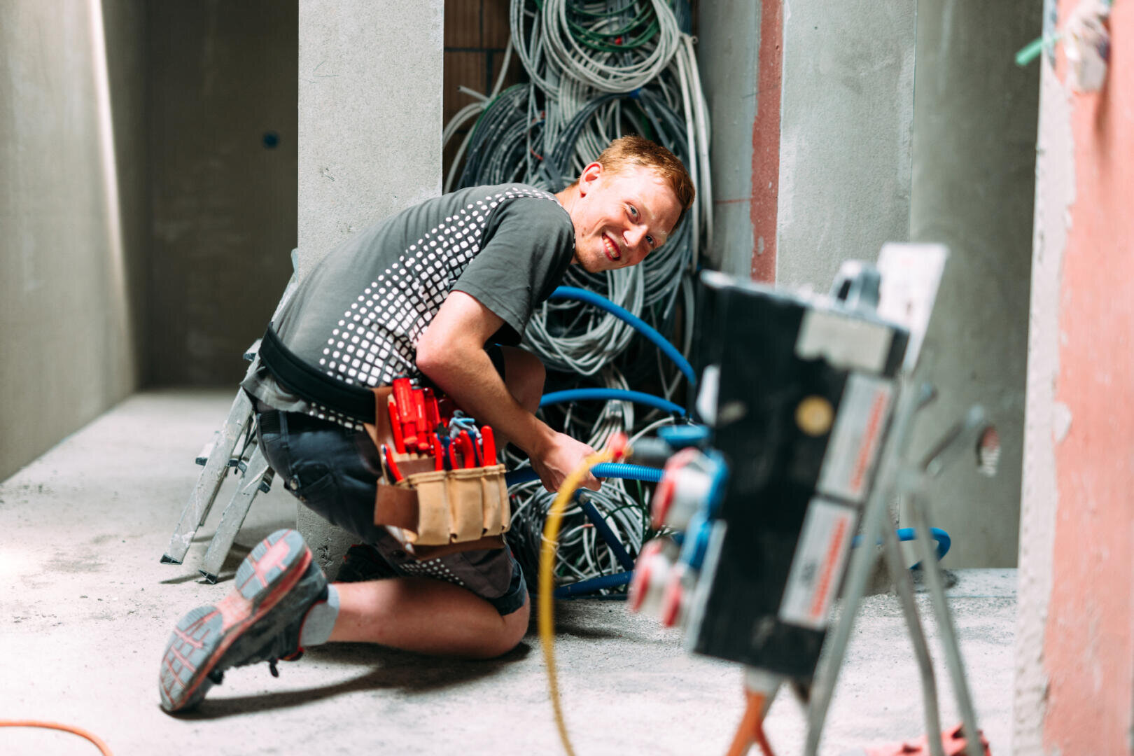 Ein lächelnder Elektriker mit einem Werkzeuggürtel kniet auf dem Boden und arbeitet mit bunten Kabeln in einem teilweise errichteten Gebäude. Um ihn herum sind freiliegende Drähte und Schalttafeln zu sehen.