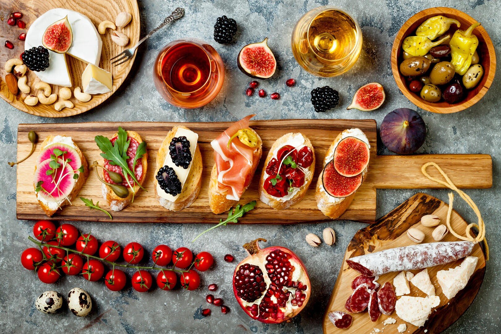 Ein rustikaler Tisch mit verschiedenen Bruschetta-Sorten, Käse, Wurstwaren, Feigen, Granatapfel, Oliven, Tomaten, Nüssen, Wachteleiern sowie Gläsern mit Rot- und Weißwein, kunstvoll angerichtet zu einem Buffet im mediterranen Stil.