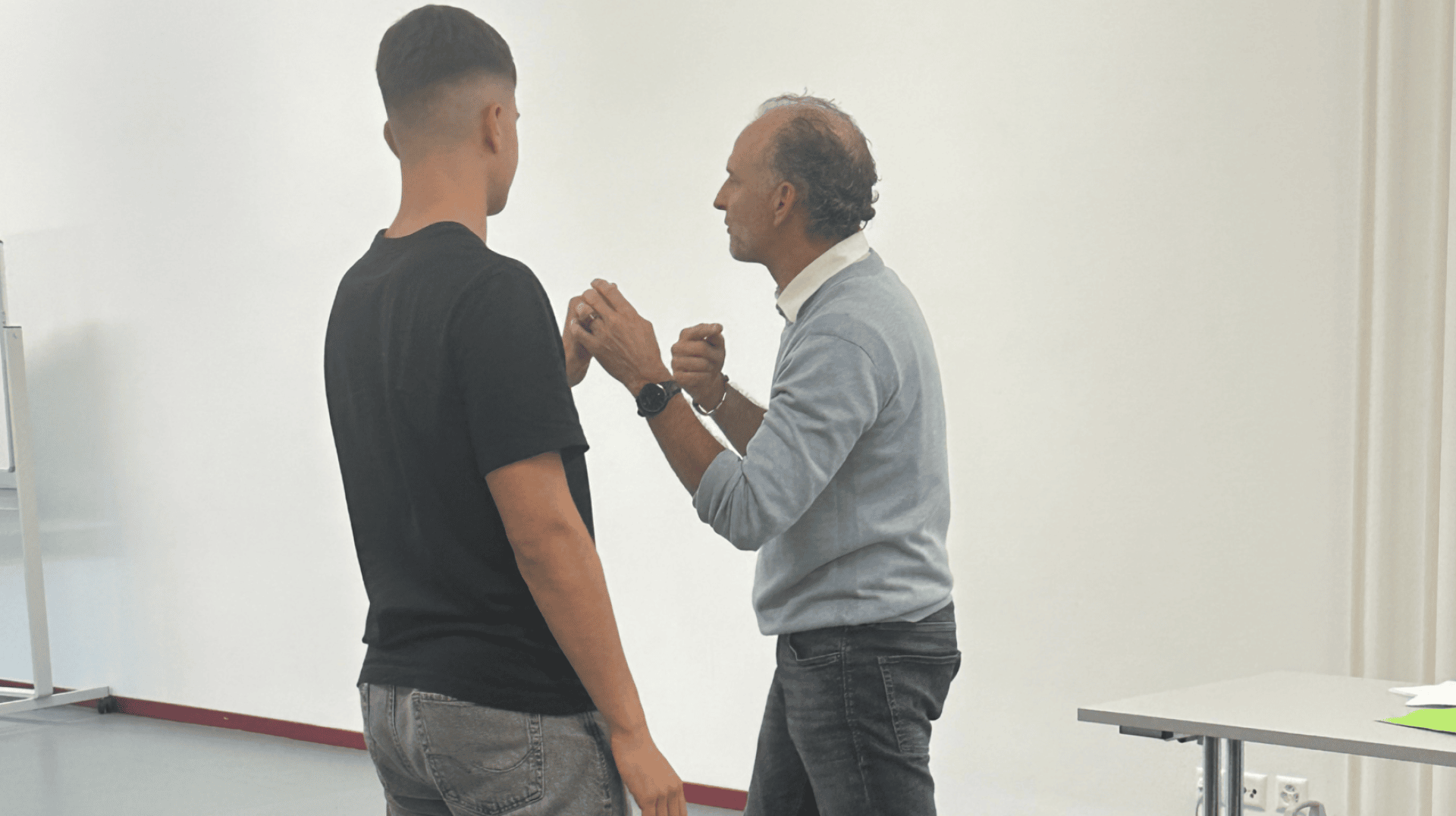 Zwei Männer stehen sich in einem Raum gegenüber. Der eine, der ein schwarzes T-Shirt und Jeans trägt, steht mit dem Rücken zur Kamera. Der andere, in einem hellen Pullover und Jeans, scheint zu sprechen oder mit erhobenen Händen zu gestikulieren.