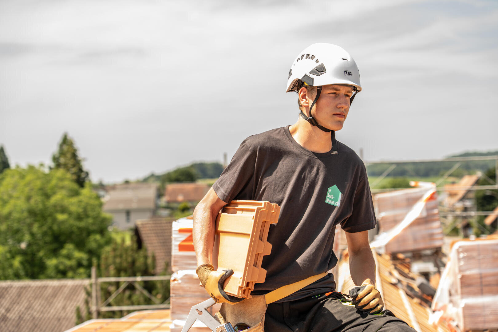 Ein Bauarbeiter mit einem weißen Helm und Handschuhen sitzt auf einem Dach und hält einen Dachziegel. Er ist mit einem dunklen T-Shirt bekleidet und blickt nachdenklich in die Ferne. Um ihn herum sind Dachmaterialien zu sehen und im Hintergrund Bäume.