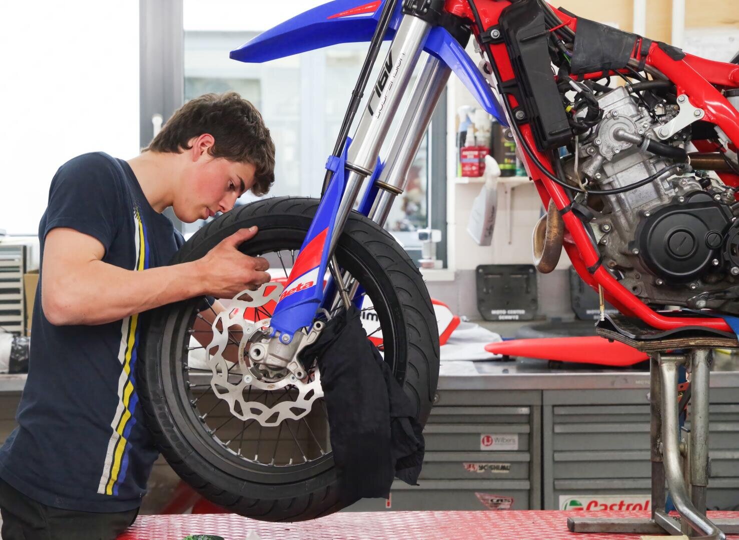 Ein junger Mann arbeitet in einer Werkstatt konzentriert am Vorderrad eines rot-blauen Motorrads und benutzt Werkzeuge, um die Bremsscheibe einzustellen. Im Hintergrund sind Werkstattschränke und Geräte zu sehen.