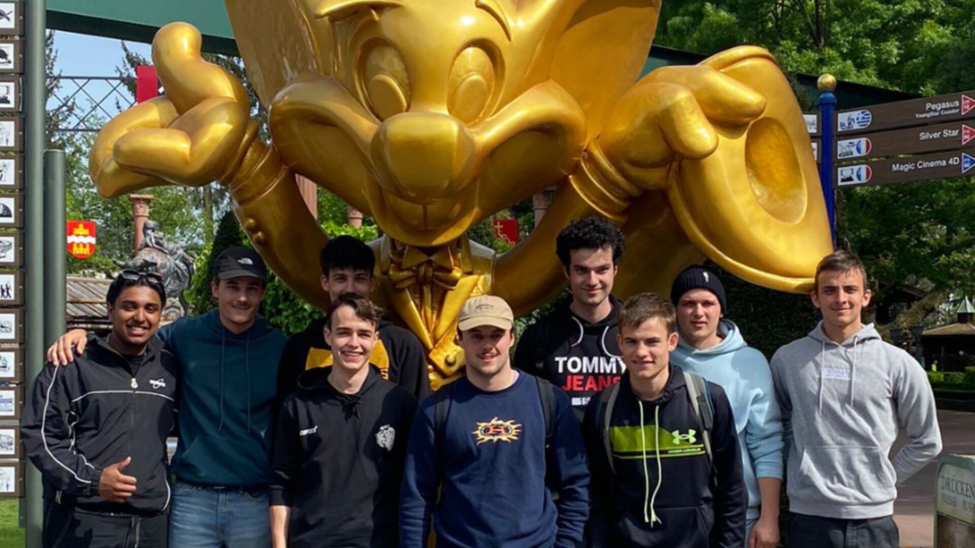 Eine Gruppe von neun jungen Männern posiert und lächelt vor einer großen goldenen Cartoon-Statue im Freien, mit Bäumen und Wegweisern im Hintergrund.