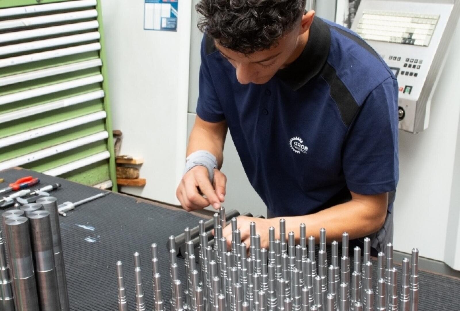 Eine Person in einem blauen Hemd prüft oder montiert Metallstangen, die auf einer Werkbank in einer Industrie- oder Fertigungsumgebung angeordnet sind, wobei im Hintergrund Werkzeuge und Geräte zu sehen sind.