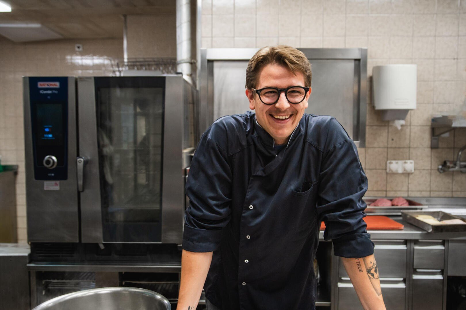 Ein lächelnder Koch mit Brille und dunkler Uniform steht in einer Profiküche und stützt sich mit den Händen auf einer Arbeitsplatte ab. Im Hintergrund sind große Geräte aus rostfreiem Stahl und geflieste Wände zu sehen.