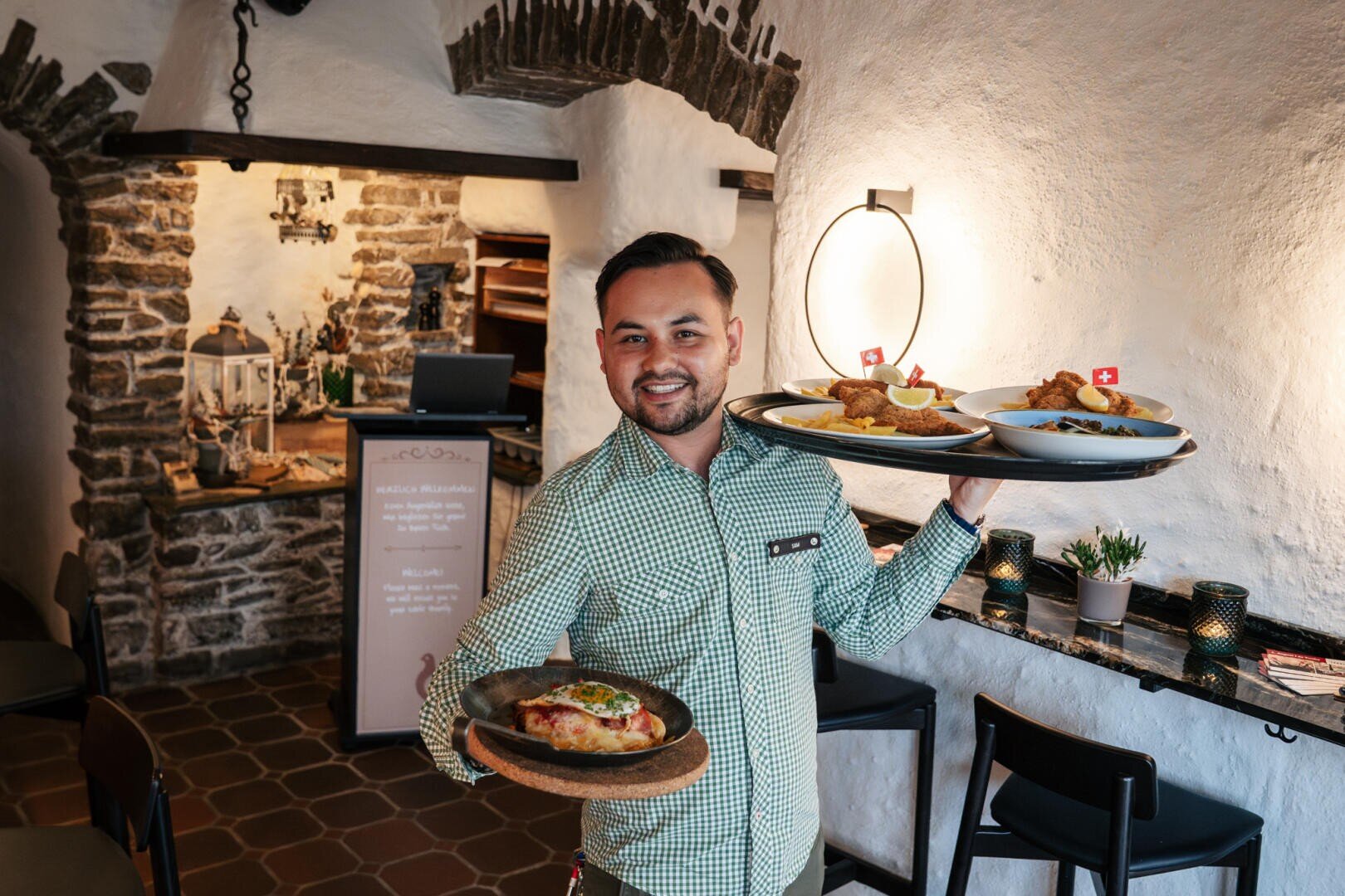 Ein lächelnder Kellner in einem grün karierten Hemd hält Teller mit Essen in einem gemütlichen Restaurant mit weißen Steinwänden, warmer Beleuchtung und rustikaler Einrichtung.