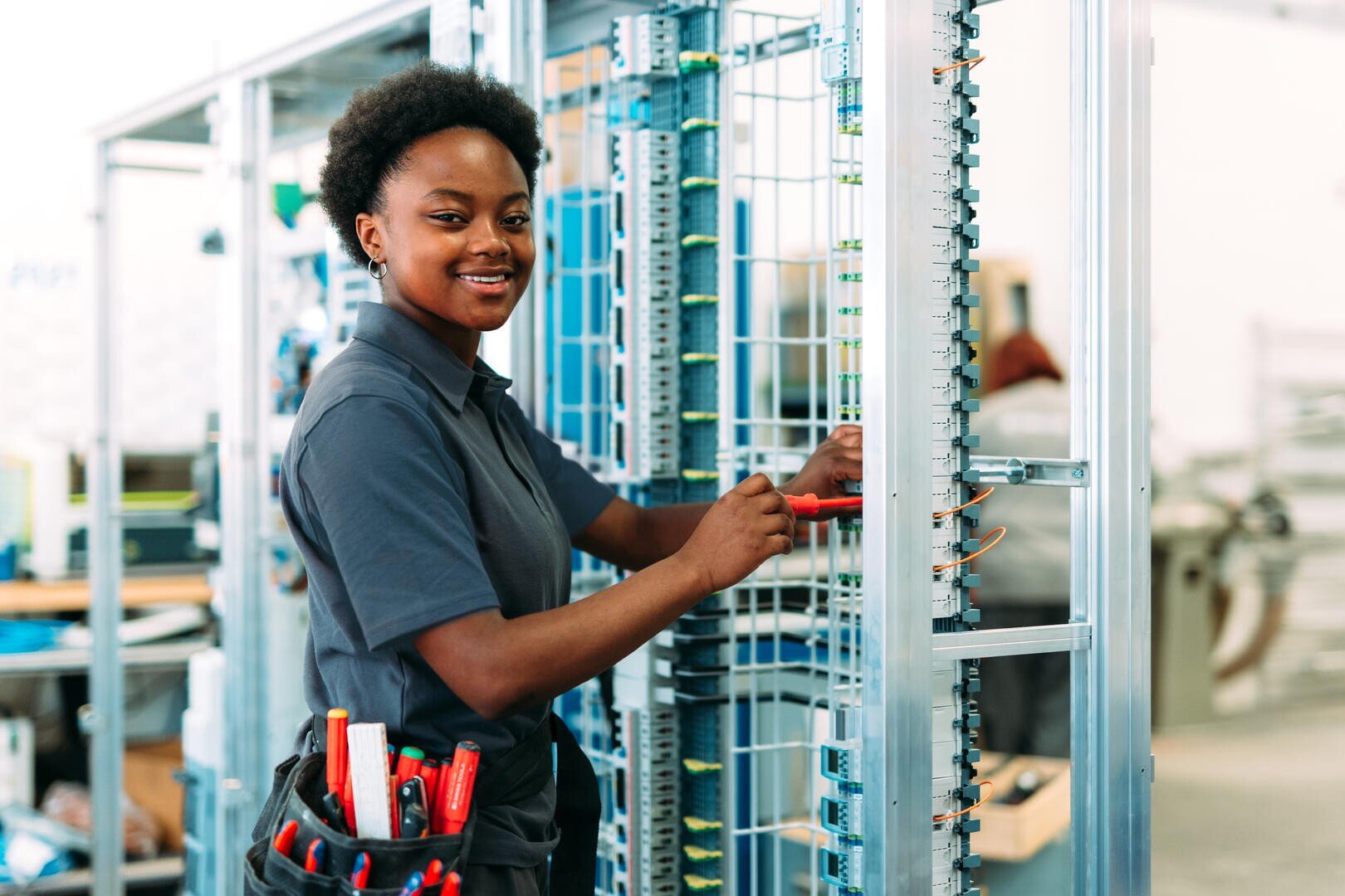 Eine lächelnde Elektrikerin in einer Arbeitsuniform arbeitet mit einem Schraubenzieher an einer Schalttafel mit elektrischen Leitungen. Sie hat einen Werkzeuggürtel mit verschiedenen Werkzeugen und steht in einer hellen, modernen Werkstatt.