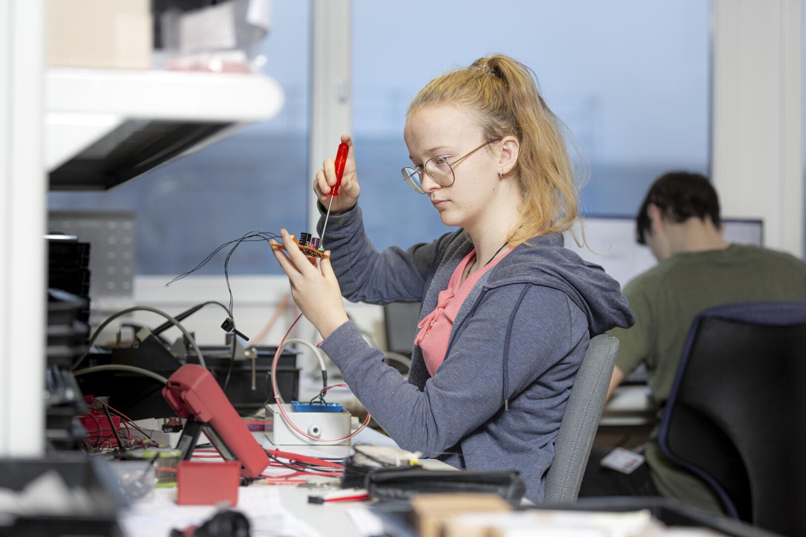 Eine junge Frau mit Brille sitzt an einem Schreibtisch und arbeitet mit einem Schraubenzieher an einem elektronischen Gerät mit Drähten. Sie ist konzentriert, umgeben von elektronischen Werkzeugen und Geräten; eine andere Person arbeitet im Hintergrund an einem Computer.