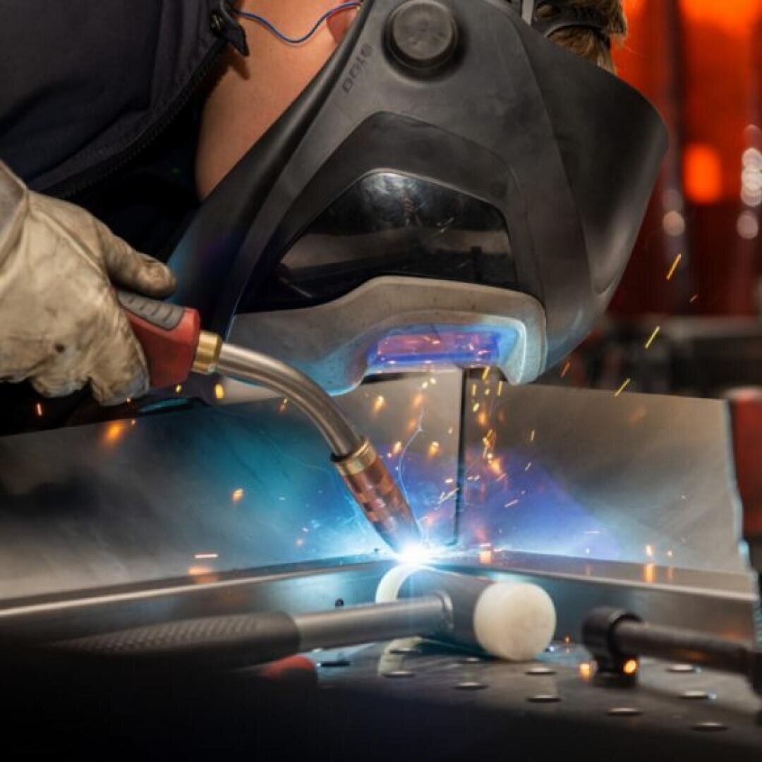 Eine Person, die einen Schweißerhelm und Handschuhe trägt, schweißt Metall und erzeugt dabei helles blaues Licht und Funken. Auf der Arbeitsfläche im Vordergrund sind verschiedene Werkzeuge, darunter ein Hammer, zu sehen.