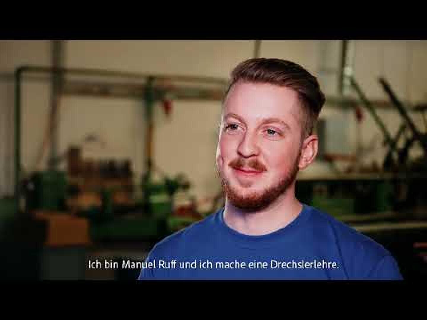 Ein junger Mann mit kurzem, hellbraunem Haar und gestutztem Bart lächelt, während er ein blaues Hemd in einer Werkstatt trägt. Der deutsche Text am unteren Rand lautet: Ich bin Manuel Ruff und ich mache eine Drechslerlehre.