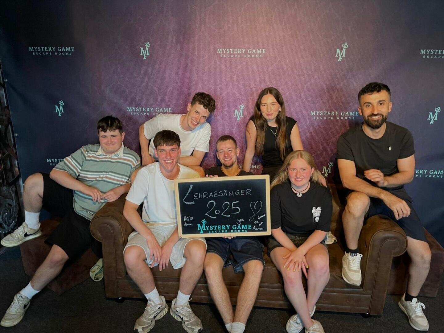 Sieben Personen sitzen und posieren auf einer braunen Couch vor einer Mystery Game Escape Room-Kulisse. Die Gruppe hält ein Kreideschild mit der Aufschrift Ehrengänger 52:25 mit Herzen und dem Mystery Game Logo.