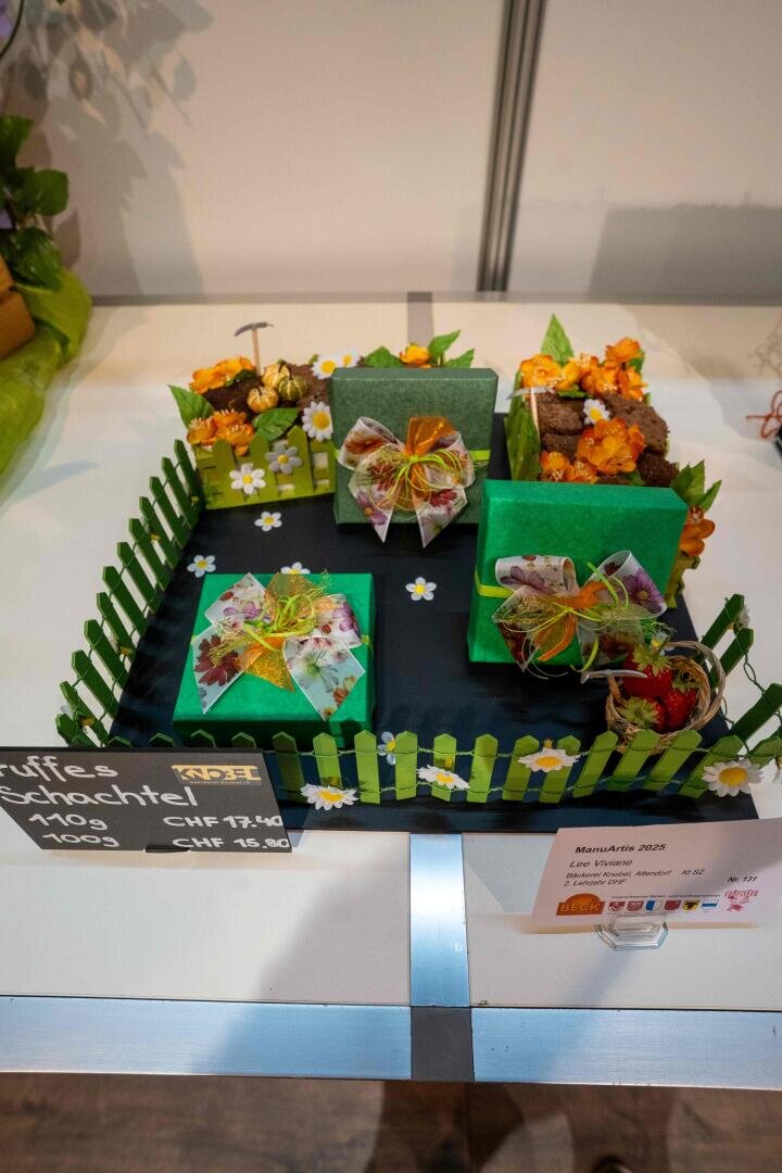 Eine dekorative Auslage mit grünen Geschenkboxen, orangefarbenen Blumen, einem kleinen Zaun und einem Sockel mit Blumenmuster, die an eine Gartenszene erinnert. Auf einer Kreidetafel sind die Preise angegeben, und auf der rechten Seite befindet sich ein Schild mit einer Aufschrift.