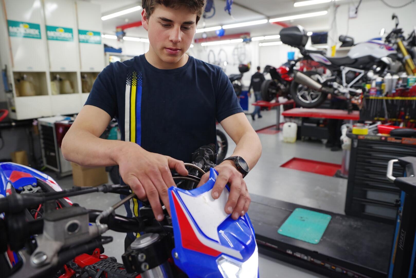 Ein junger Mann in einem marineblauen T-Shirt arbeitet in einer gut beleuchteten Werkstatt an einem blau-roten Motorrad, während im Hintergrund Werkzeuge und andere Motorräder zu sehen sind.