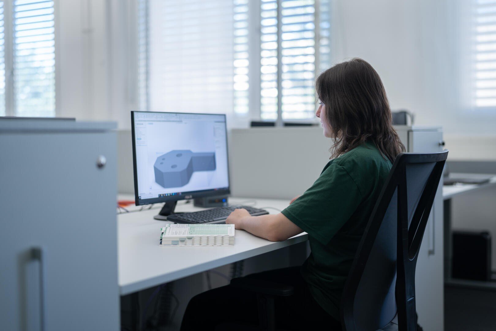 Eine Person sitzt an einem Schreibtisch und arbeitet an einem 3D-Modell, das auf einem Computermonitor in einer Büroumgebung angezeigt wird, während in der Nähe ein Notebook geöffnet ist und Licht durch die Jalousien einfällt.