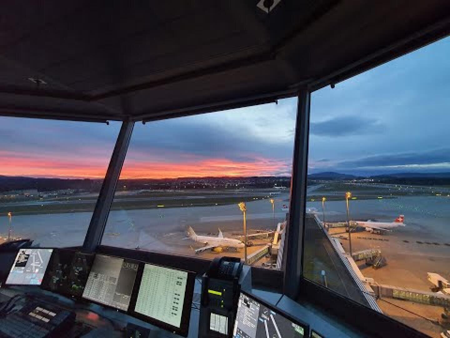 Blick auf einen Flughafenkontrollturm bei Sonnenuntergang mit Blick auf Flugzeuge, die an Flugsteigen auf dem Rollfeld geparkt sind, mit Computerbildschirmen im Vordergrund und einem farbenfrohen Himmel im Hintergrund.