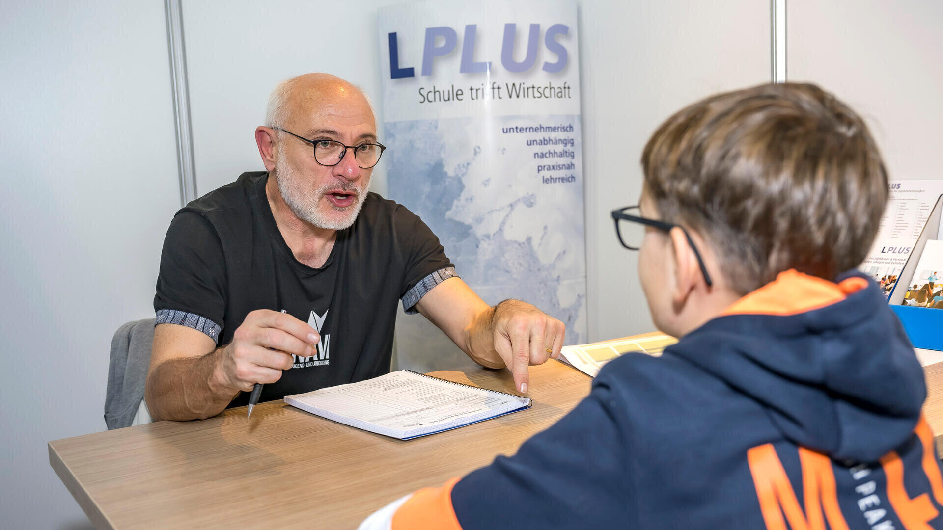 Ein älterer Mann mit Brille unterhält sich mit einem Jungen, der ihm gegenüber am Schreibtisch sitzt. Auf dem Tisch liegen Papiere und ein Stift. Auf einem Banner hinter den beiden steht „LPLUS Schule fit für Wirtschaft“.