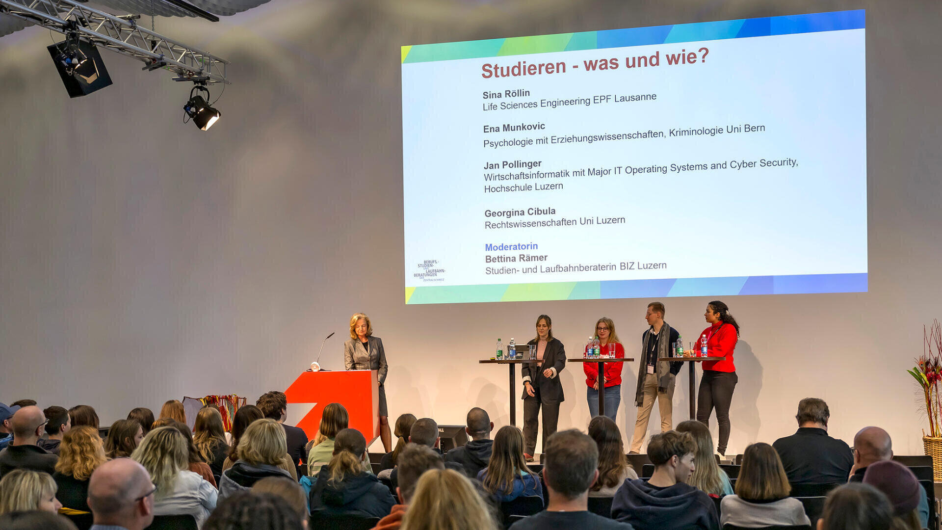 Ein fünfköpfiges Gremium steht auf einer Bühne vor einem sitzenden Publikum, hinter dem eine große Präsentationsfolie mit dem Titel "Studieren - was und wie?" projiziert wird. Eine Person spricht von einem Podium aus, während die anderen in der Nähe warten.