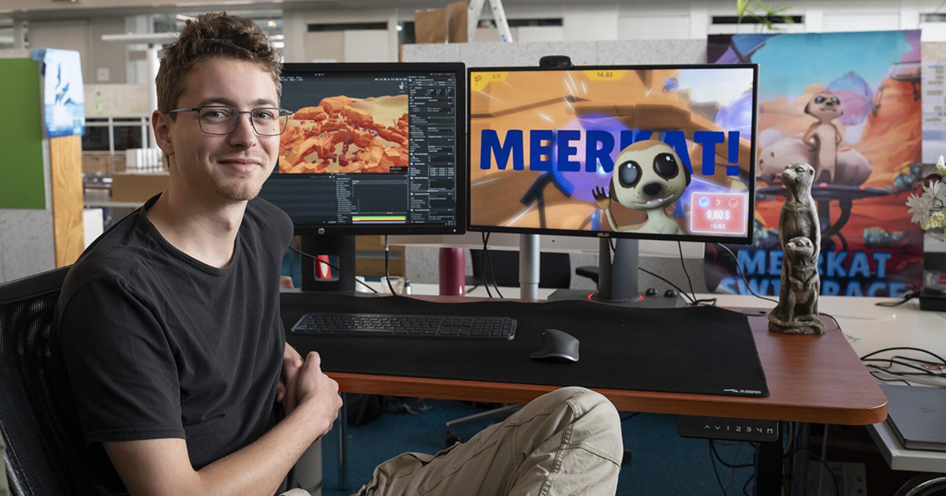 Eine Person sitzt an einem Schreibtisch mit einem Computer, der animierte Erdmännchen und ein 3D-Modellierungsprogramm zeigt. Auf dem Schreibtisch steht eine Erdmännchen-Figur, und im Hintergrund ist ein großes Poster mit MEERKAT zu sehen. Die Person lächelt in die Kamera.