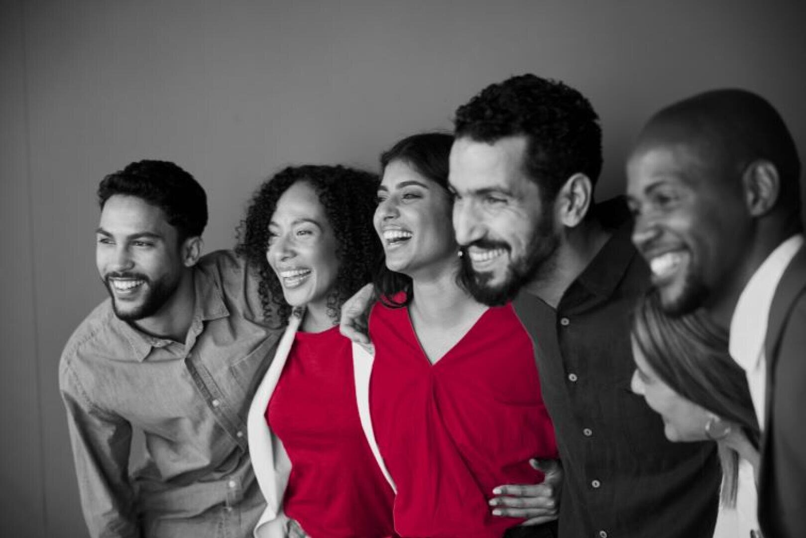 Fünf Menschen stehen dicht beieinander, lächeln und lachen. Das Bild ist in Schwarz-Weiß gehalten, mit Ausnahme von zwei Personen, deren rote Hemden farblich hervorstechen.