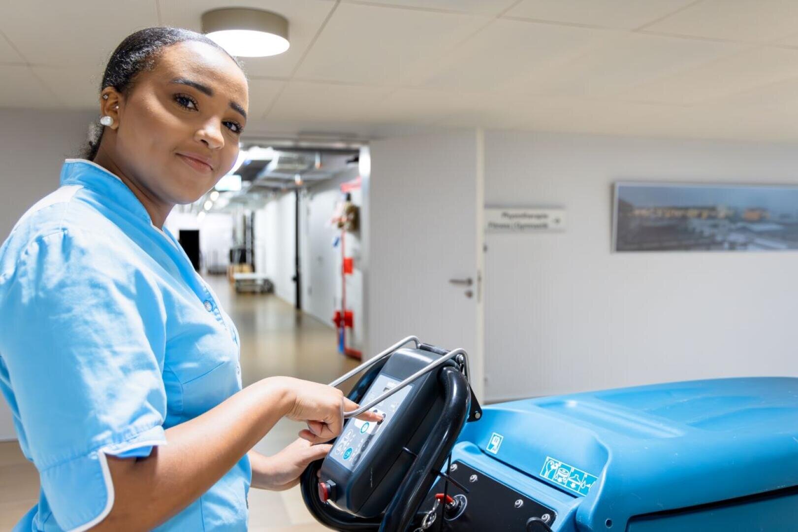 Eine Frau in einer blauen Uniform bedient in einem hellen Flur eine Reinigungsmaschine, schaut in die Kamera und lächelt leicht. An den weißen Wänden hinter ihr sind Schilder und ein Foto zu sehen.