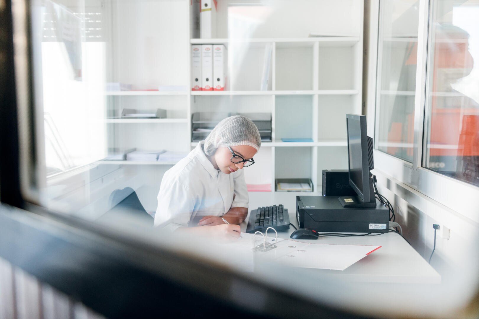 Eine Person mit Haarnetz und Brille sitzt an einem Schreibtisch in einem Büro und schreibt auf Papieren neben einem Computer und einem Ordner, gesehen durch ein Fenster. Im Hintergrund befinden sich Regale mit Akten.