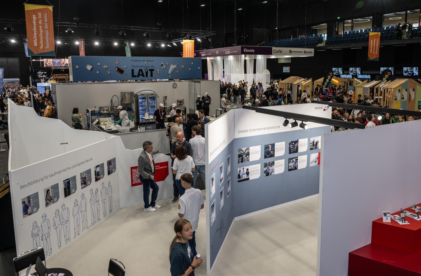 Eine belebte ĂŒberdachte Ausstellungshalle, in der die Menschen zwischen weiĂen AusstellungsstĂ€nden und Informationstafeln mit verschiedenen StĂ€nden, Bannern und interaktiven Bereichen umhergehen. Die AtmosphĂ€re ist lebendig und einnehmend.
