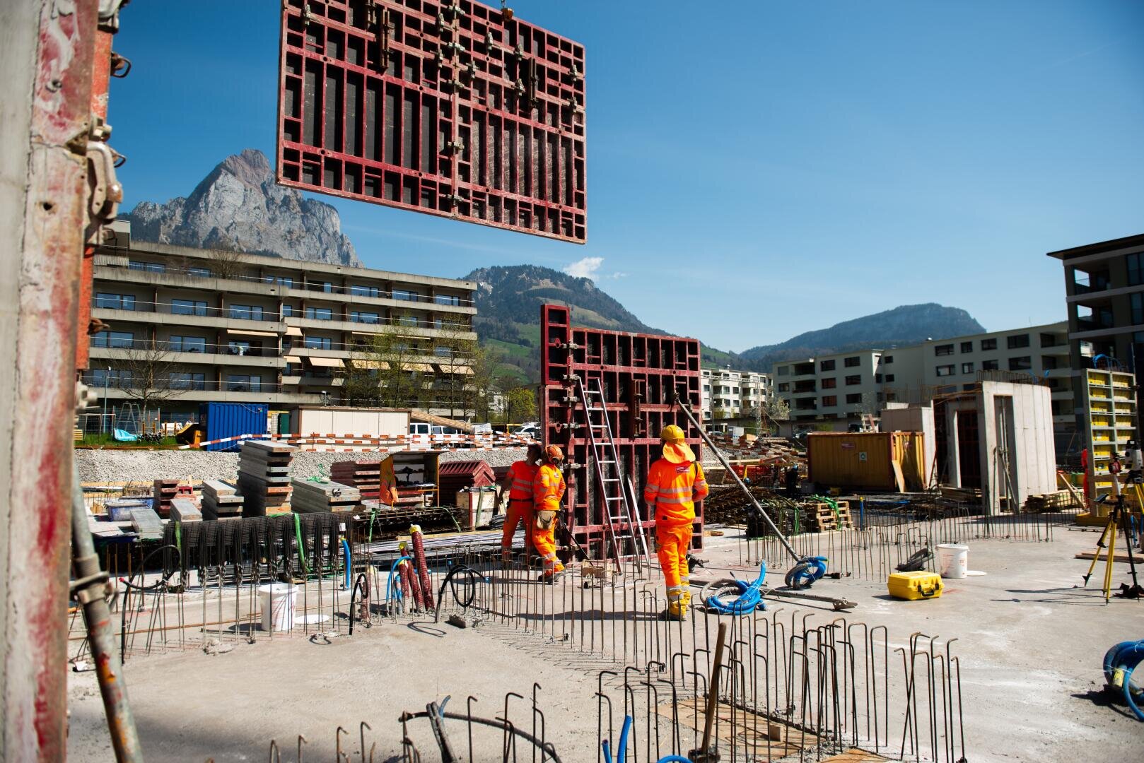 Zwei Bauarbeiter in orangefarbener Kleidung stehen auf einer Baustelle mit freiliegendem Betonstahl. Eine große Metallplatte wird gerade von einem Kran hochgezogen. Im Hintergrund sind Wohnhäuser und Berge unter einem klaren blauen Himmel zu sehen.