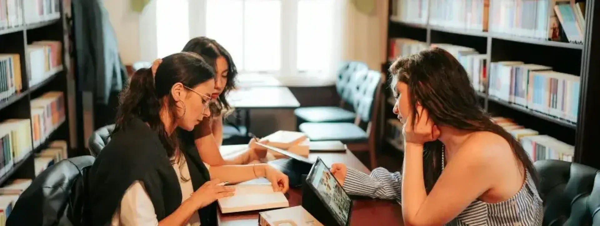 Drei Frauen sitzen an einem Tisch in einer Bibliothek, lesen und diskutieren über Bücher. An den Wänden stehen Regale voller Bücher, und durch ein Fenster hinter ihnen fällt das Sonnenlicht ein.