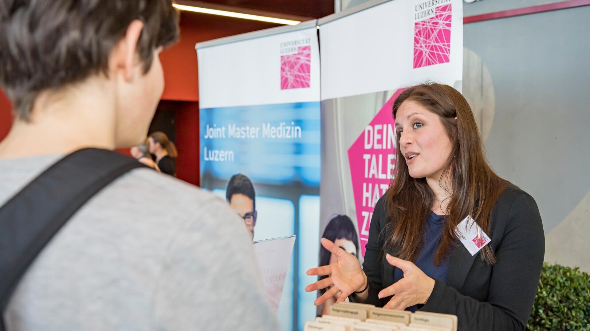 Eine Frau an einem Informationsstand spricht mit einer Besucherin. Hinter ihr werben Plakate für einen Masterstudiengang an der Universität Luzern. Sie gestikuliert mit den Händen, während sie ein Gespräch führt.