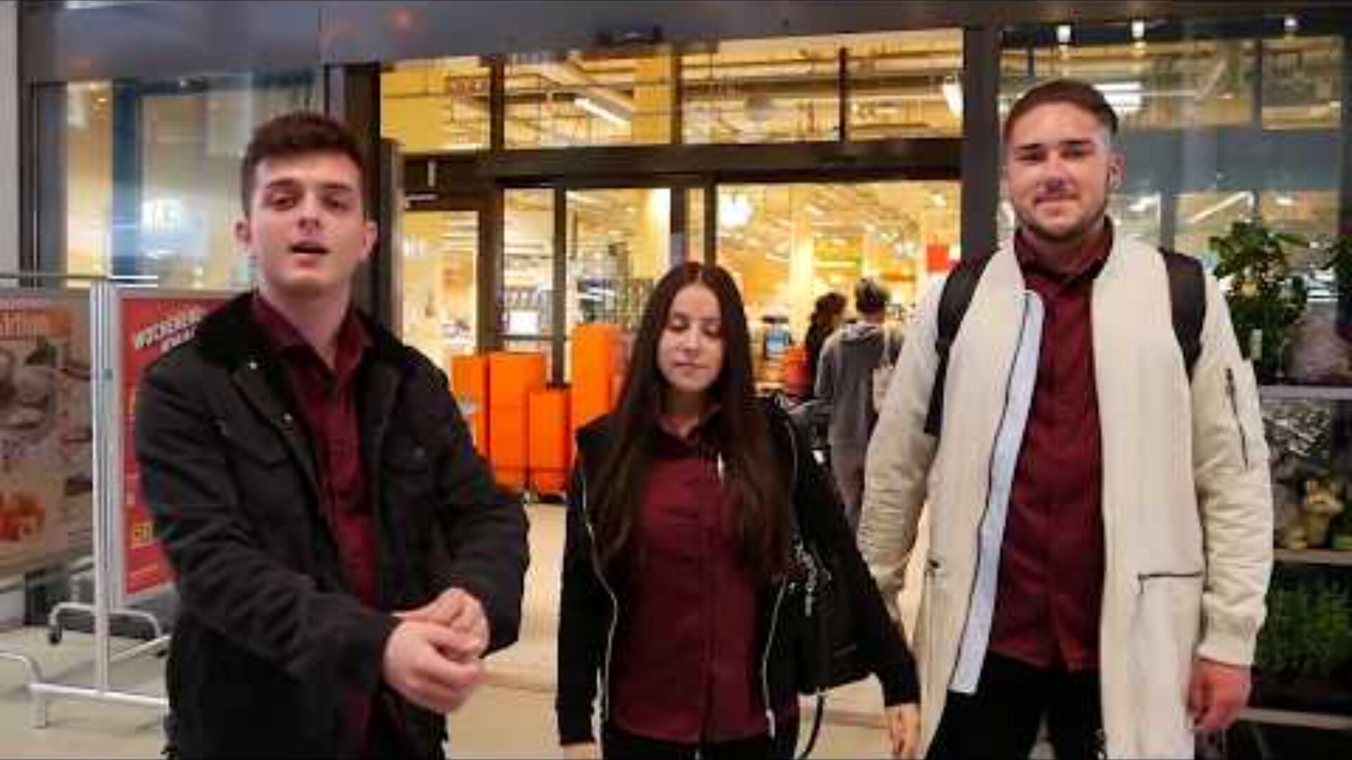 Drei junge Erwachsene, zwei Männer und eine Frau, stehen vor einem gut beleuchteten Ladeneingang. Sie tragen alle kastanienbraune Hemden und Jacken, wobei die Frau in der Mitte eine schwarze Handtasche in der Hand hält.