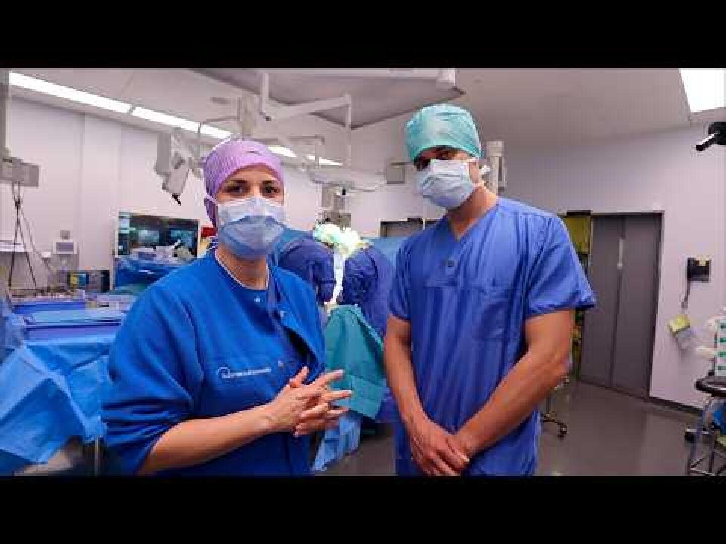 Zwei medizinische Fachkräfte, die blaue OP-Kleidung, Masken und Hauben tragen, stehen in einem gut beleuchteten Operationssaal. Im Hintergrund sind chirurgische Instrumente und weiteres Personal zu sehen.
