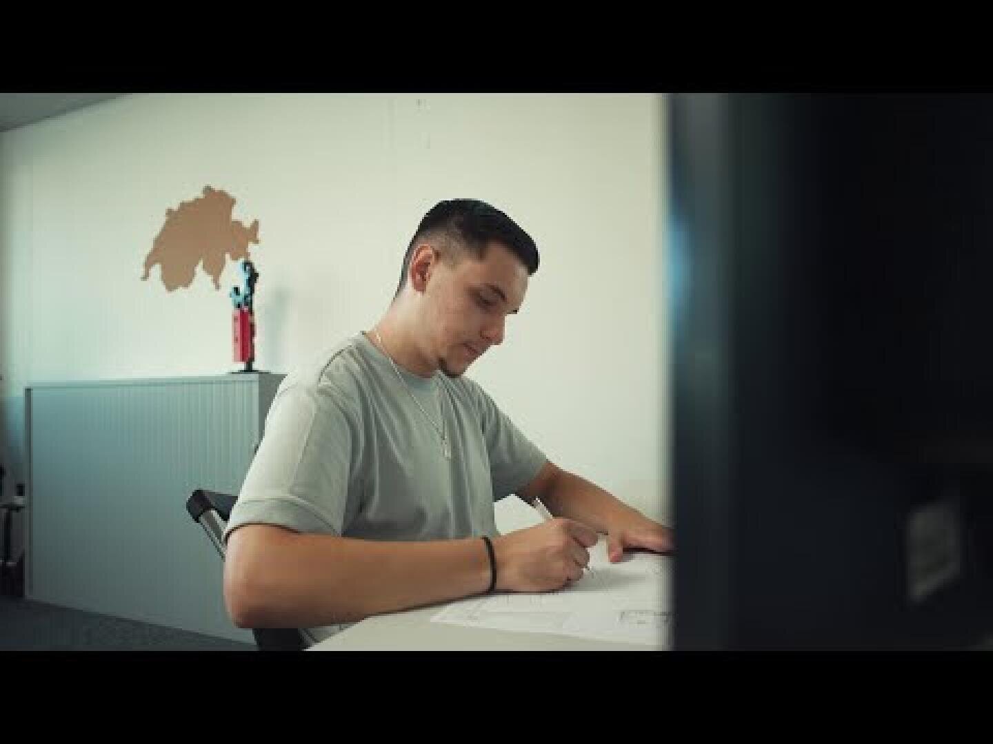 Ein Mann sitzt an einem Schreibtisch und schreibt in einem hellen Büro auf Papier. An der Wand hinter ihm ist eine Karte oder ein Kunstwerk angebracht, daneben ein roter Feuerlöscher.