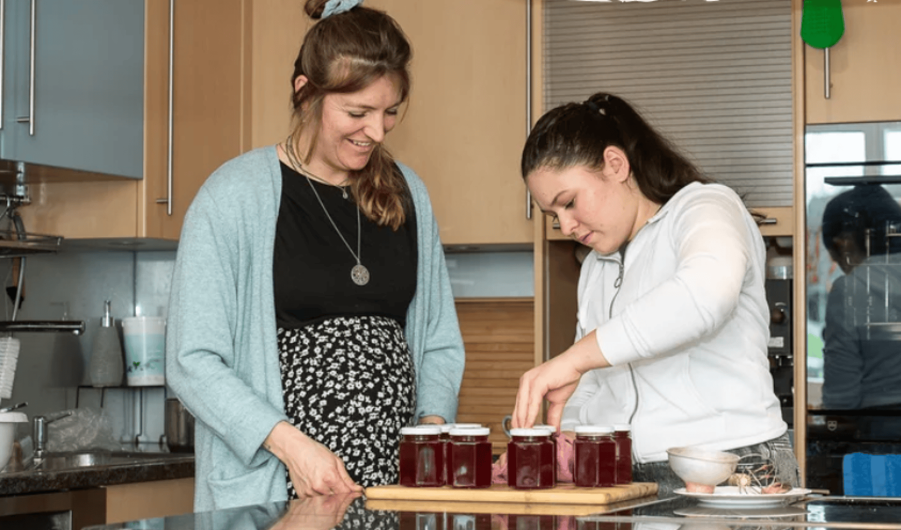 Zwei Frauen in einer Küche, die lächelnd Deckel auf Gläser mit roter Marmelade setzen, die auf einem Holzbrett stehen. Im Hintergrund der Küche sind Schränke, eine Spüle und verschiedene Küchenutensilien zu sehen.