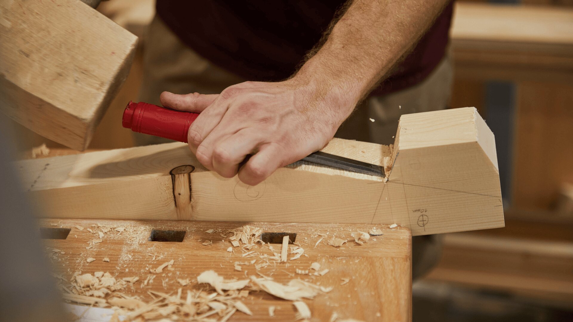 Eine Person schnitzt mit einem Meißel mit rotem Griff und einem Holzhammer eine präzise Fuge in ein Stück helles Holz, wobei die Holzspäne auf der Werkbank verstreut sind.