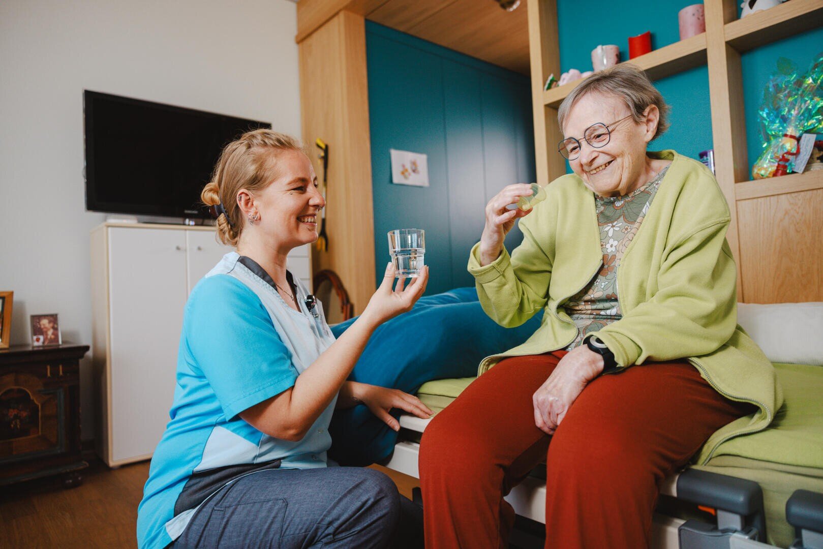 Eine Pflegekraft lächelt, während sie einer fröhlichen älteren Frau, die auf einem Bett in einem gemütlichen, hell erleuchteten Zimmer sitzt, ein Glas Wasser und Medikamente reicht.