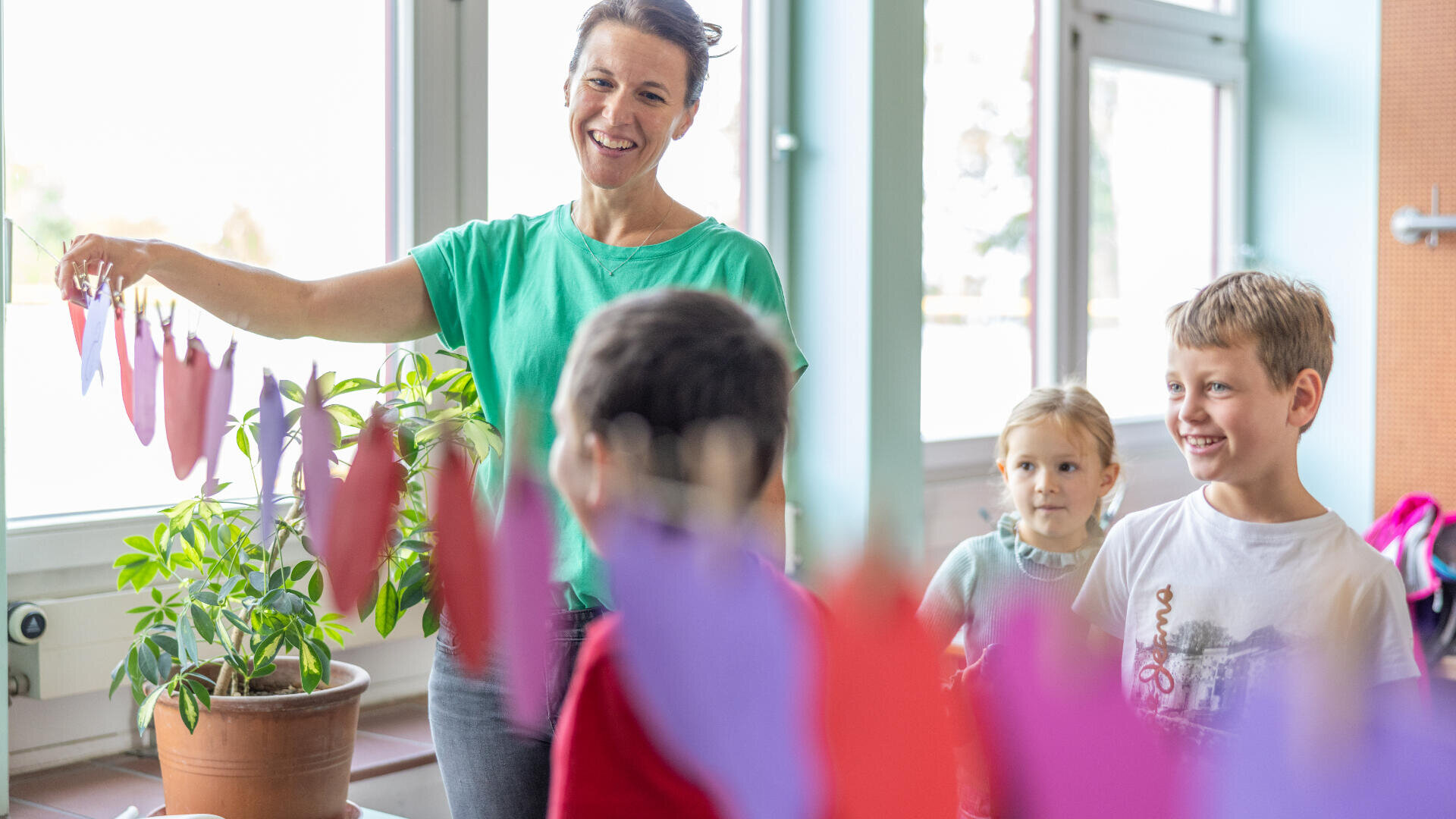 Eine Frau lächelt, während sie eine Schnur mit bunten Papierformen hält. Drei kleine Kinder stehen in der Nähe, beobachten sie und lächeln. Sie befinden sich in einem Innenraum in der Nähe großer Fenster, durch die Sonnenlicht einfällt.