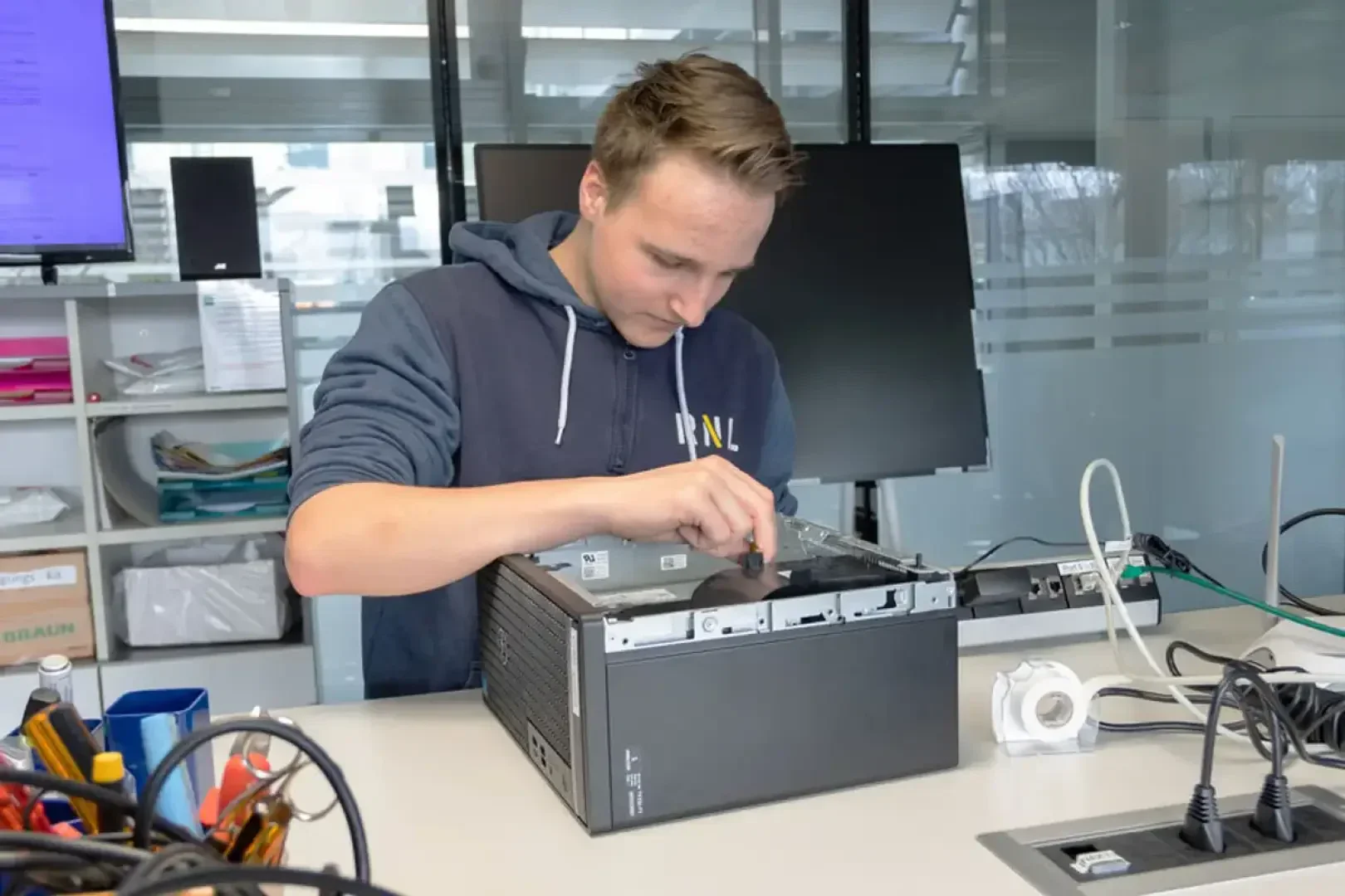 Eine Person in einem Kapuzenpulli repariert oder baut einen Desktop-Computer auf einem Tisch zusammen, umgeben von Werkzeugen und elektronischen Geräten in einer modernen Büro- oder Laborumgebung.