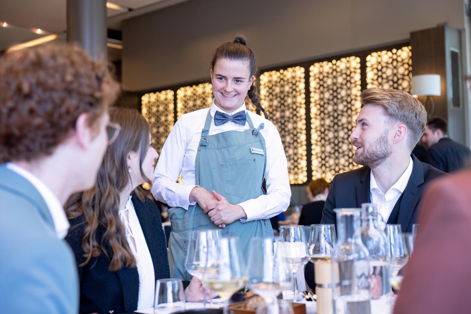 Ein lächelnder Kellner in blauer Schürze und Fliege steht an einem Tisch und unterhält sich mit vier Gästen in einem modernen, gut beleuchteten Restaurant. Weingläser und Flaschen stehen auf dem Tisch.