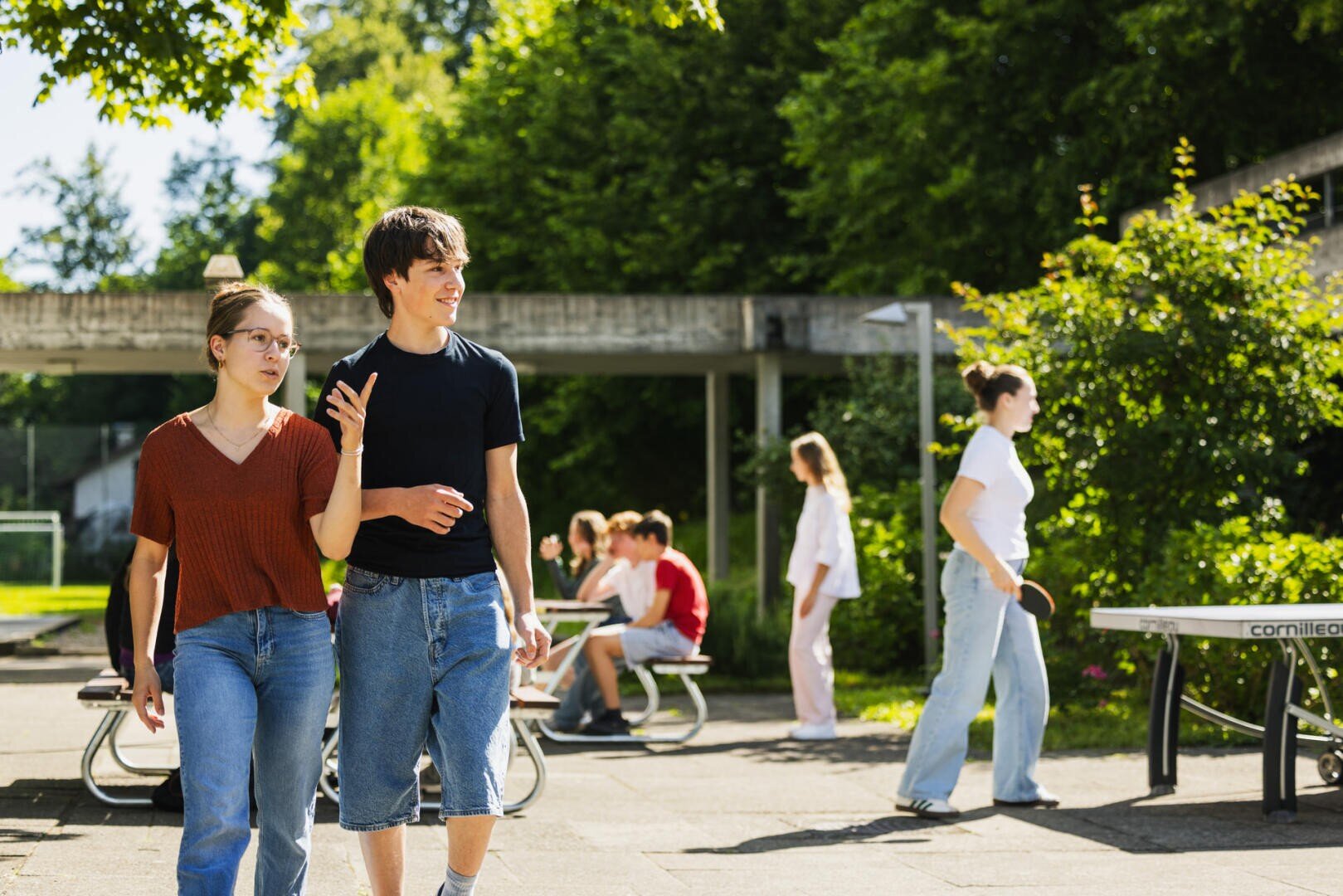 Eine junge Frau und ein junger Mann spazieren gemeinsam auf einem Schulgelände im Freien, während im Hintergrund andere an Picknicktischen sitzen.
