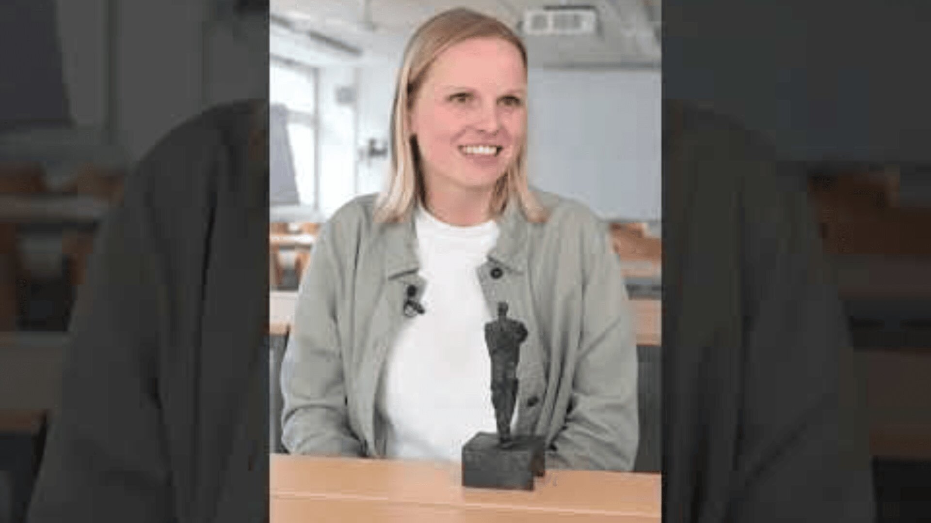 Eine Frau mit blondem Haar, die eine helle Jacke und ein weißes Hemd trägt, sitzt an einem Schreibtisch in einem Klassenzimmer und lächelt. Vor ihr steht eine kleine schwarze Statue einer stehenden Figur.