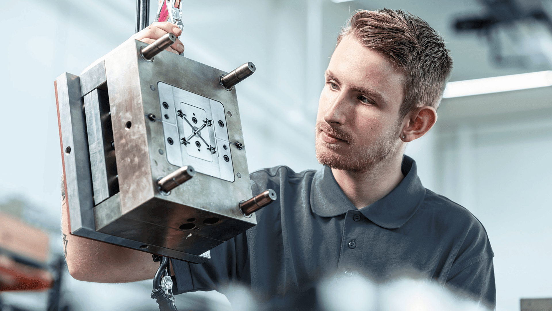 Ein junger Mann in einem grauen Polohemd prüft ein Metallformwerkzeug, das er in einer gut beleuchteten Werkstatt oder Produktionsumgebung vorsichtig mit beiden Händen hochhält.
