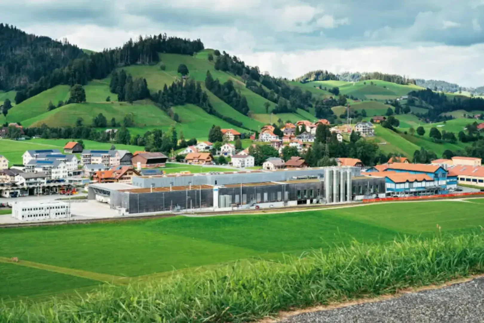 In einem grünen Tal steht ein modernes Industriegebäude, umgeben von Hügeln, traditionellen Häusern mit roten Dächern und vereinzelten Bäumen unter einem bewölkten Himmel.
