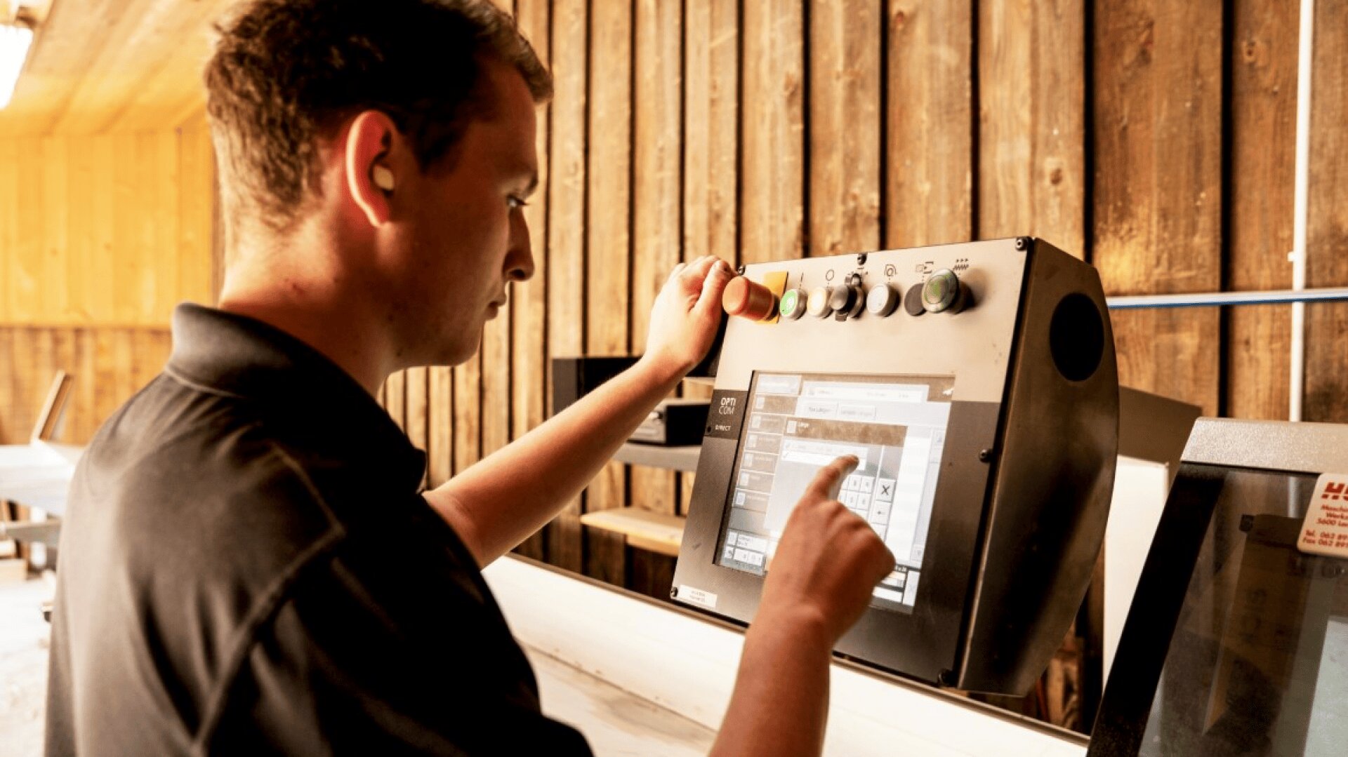 Ein Mann in einem schwarzen Hemd bedient ein Touchscreen-Bedienfeld in einer Industrieumgebung mit Holzwänden im Hintergrund. Mit einer Hand berührt er den Bildschirm, mit der anderen drückt er eine Taste.