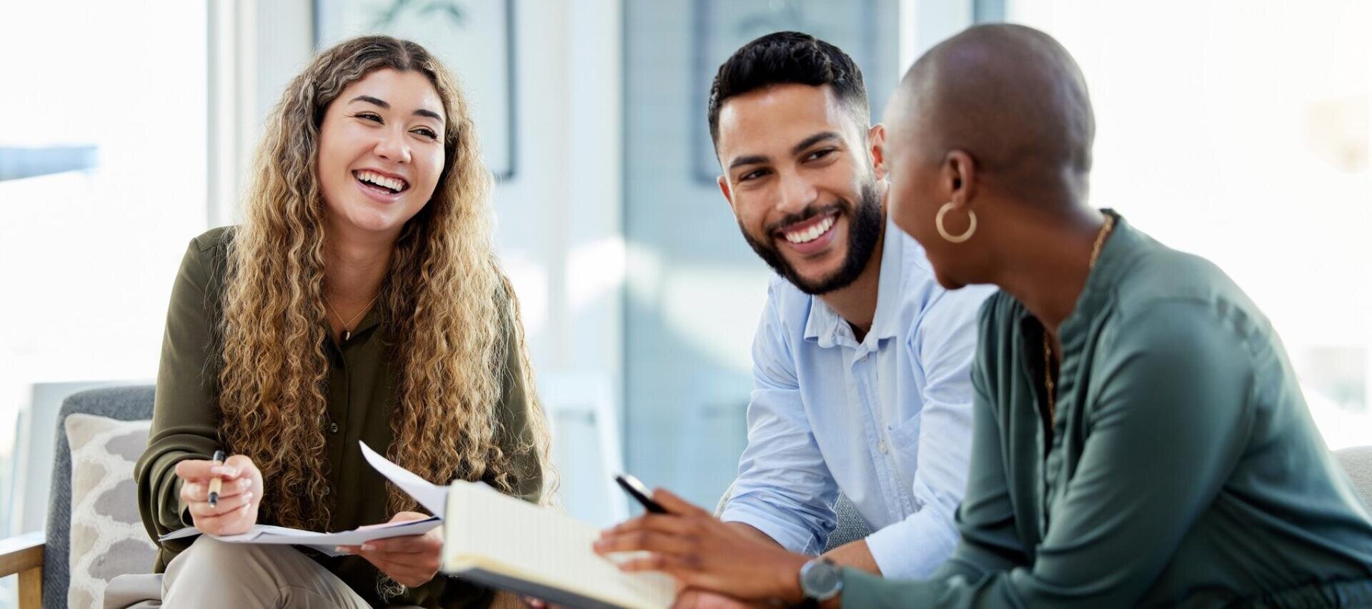 Drei Personen sitzen zusammen, lächeln und unterhalten sich, während sie Notizbücher und Stifte in der Hand halten – in einem hellen, modernen Büro.