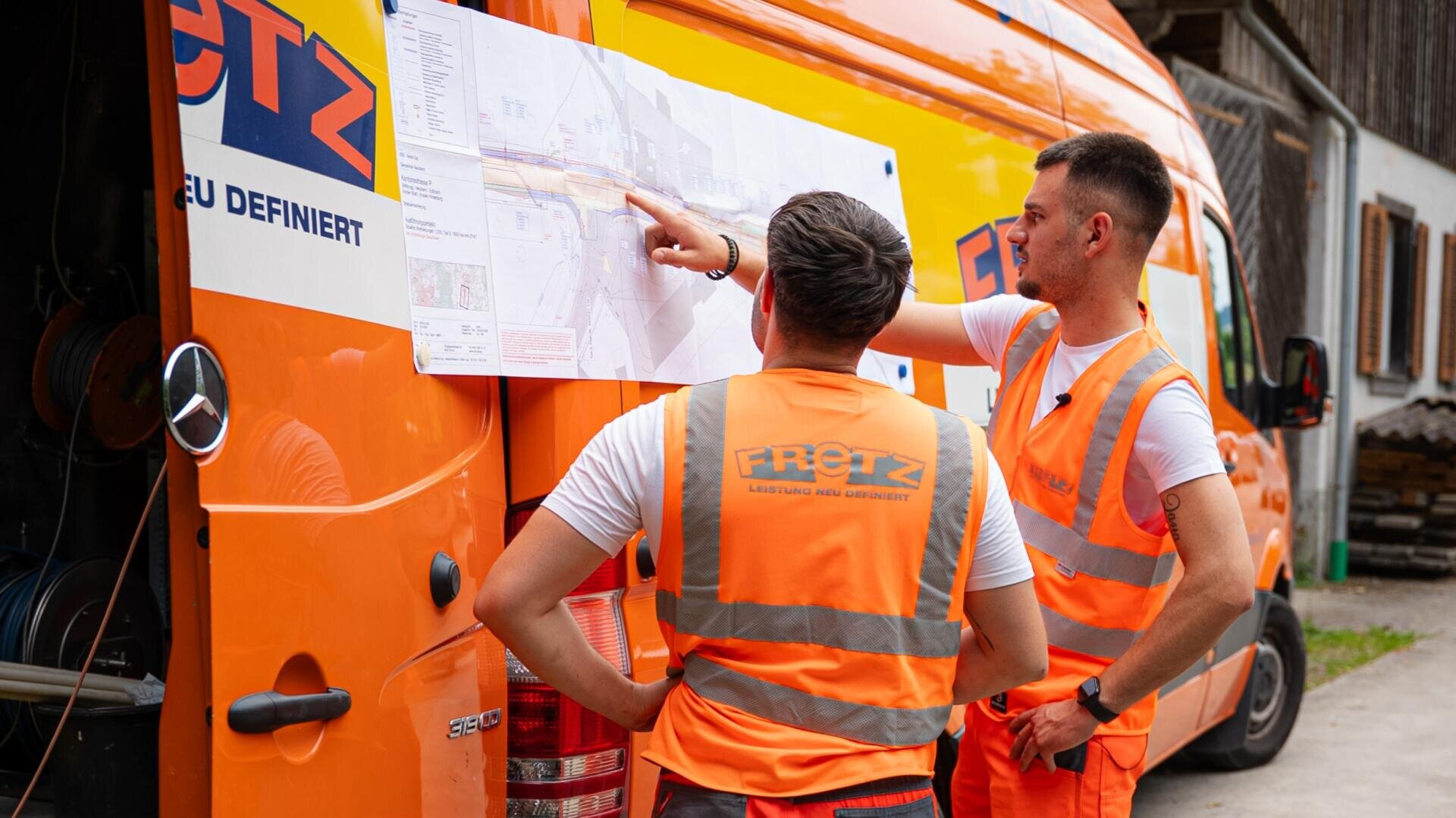 Zwei Arbeiter in orangefarbenen Sicherheitswesten betrachten eine große Karte, die an der Seite eines orangefarbenen Lieferwagens befestigt ist, und besprechen ihre Pläne. Der Lieferwagen und die Kleidung tragen das Fretz-Logo. Sie befinden sich im Freien in der Nähe von Gebäuden.