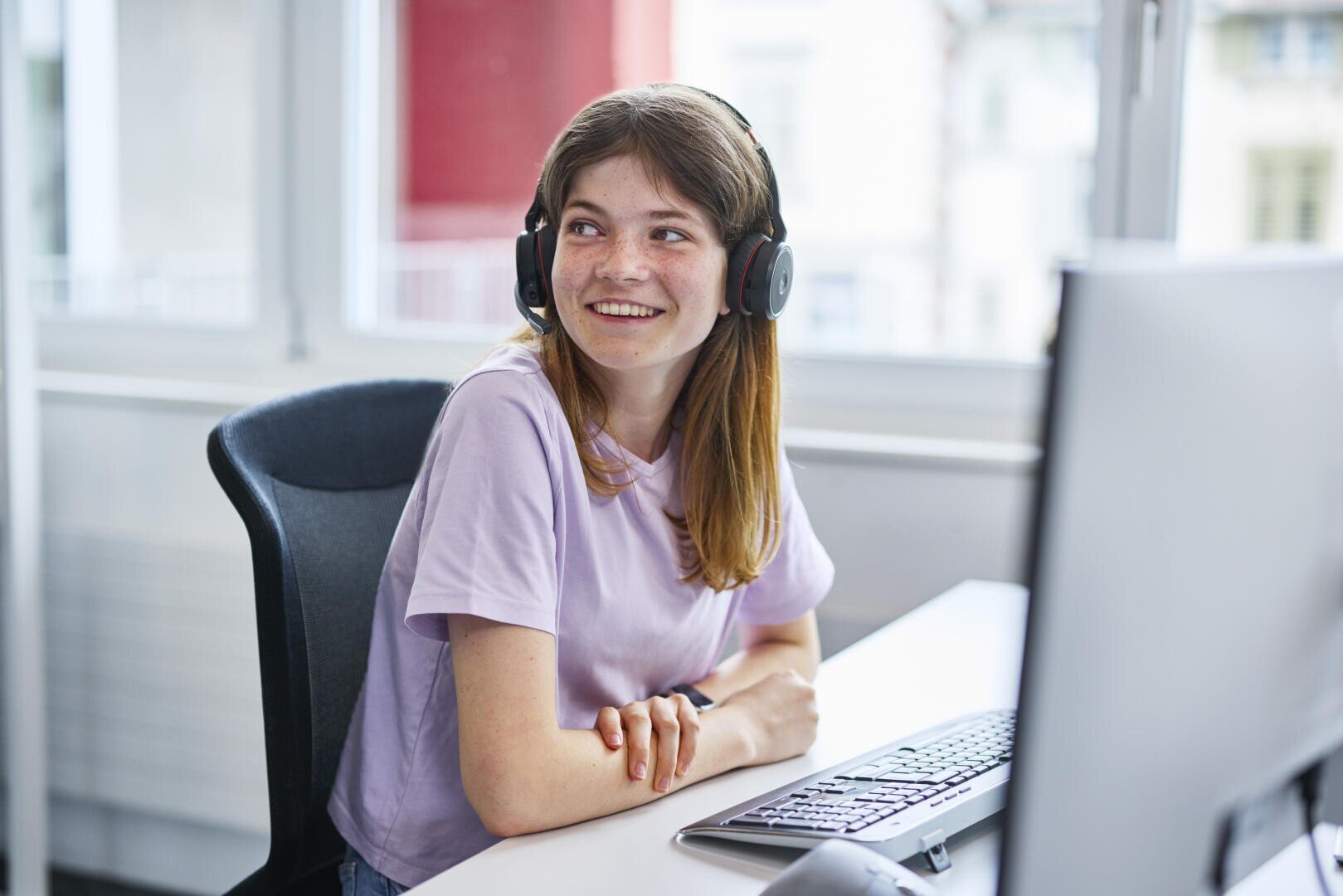 Eine junge Frau, die Kopfhörer und ein lavendelfarbenes Hemd trägt, lächelt, während sie an einem Schreibtisch mit Computer, Tastatur und Maus in einem hellen Büroraum sitzt.
